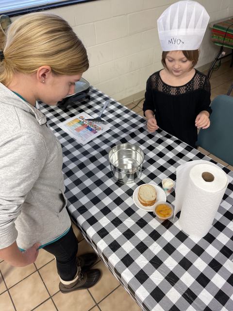 Two children at checkered table with chef hat reading "MYO", sandwich and metal pail.