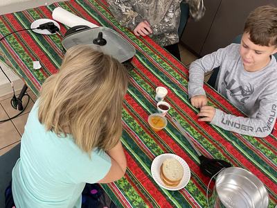 Three children seated at a striped tablecloth with a sandwich, condiments, and a cooking pan