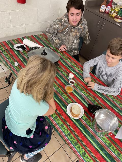 Three children seated at a striped tablecloth with a sandwich, condiments, and a cooking pan