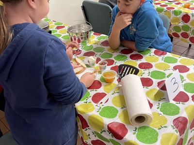 Children at a table with apple-patterned cloth preparing food; table number 3 visible