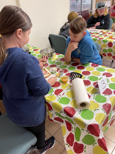 Children at a table with apple-patterned cloth preparing food; table number 3 visible