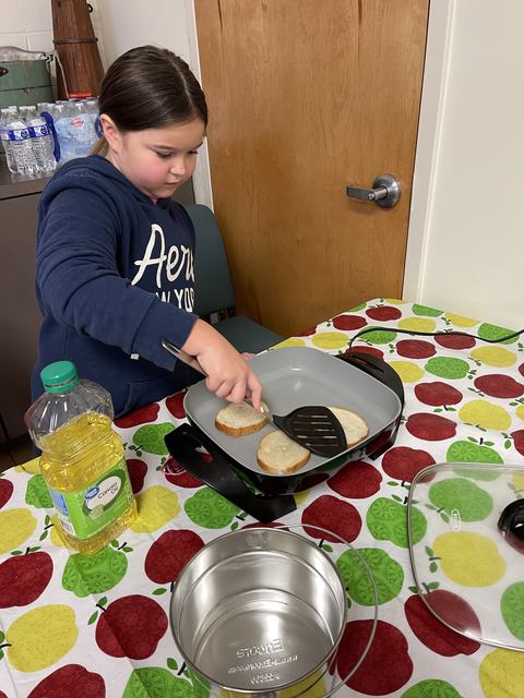 Child using spatula to cook bread slices in electric skillet on apple-patterned tablecloth