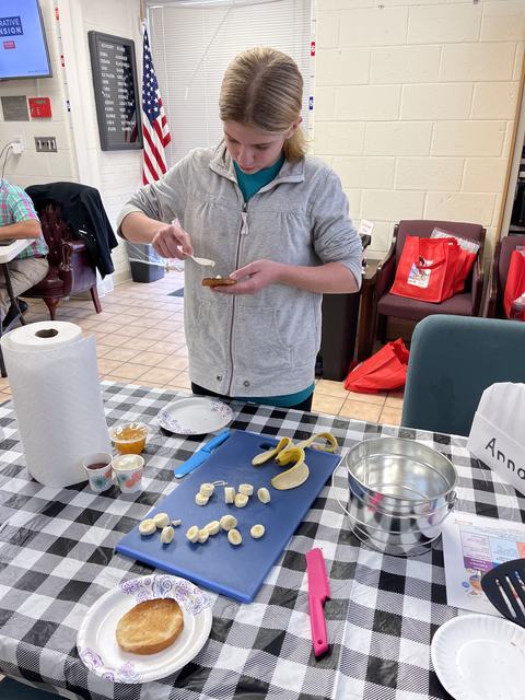 Person spreading topping on a toasted bun at a table with sliced bananas and utensils