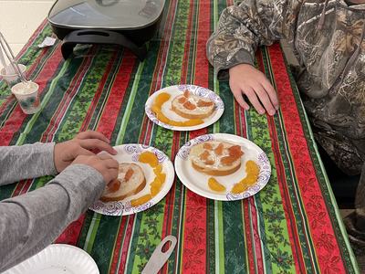 Two people assembling bagels with cream cheese and mandarin slices on holiday tablecloth