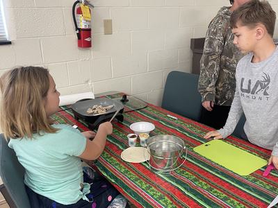 Children at table cooking chicken on electric griddle with tortillas, bowls, and cutting board