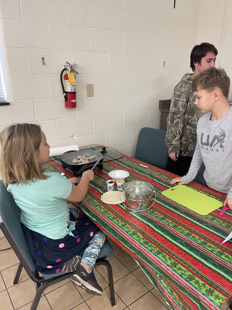 Children at table cooking chicken on electric griddle with tortillas, bowls, and cutting board