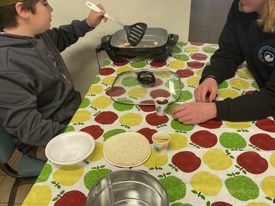 Two people at a table cooking chicken on an electric griddle with tortillas and cups nearby