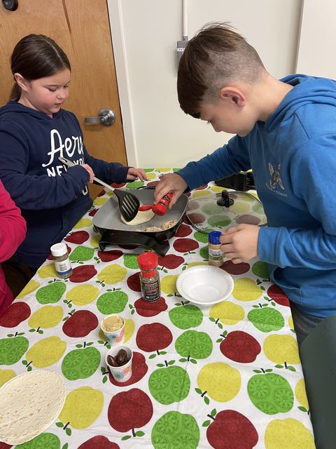 Two children at table seasoning and flipping food on an electric griddle