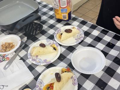 Three tortillas on paper plates topped with toothpicked dates; hand reaching into shredded-cheese bag