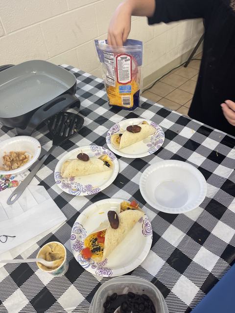 Three tortillas on paper plates topped with toothpicked dates; hand reaching into shredded-cheese bag