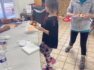 Two children holding plates with hot dogs; child on right wearing chef hat labeled "Anna"