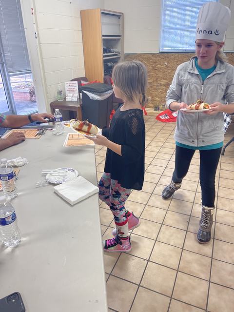 Two children holding plates with hot dogs; child on right wearing chef hat labeled "Anna"
