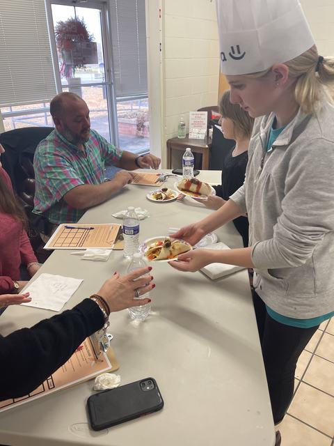 Girl in paper chef hat serving a plate of food to seated people at a judging table