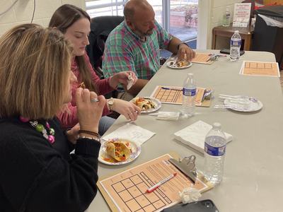 Three people seated at a table eating from paper plates, clipboards and water bottles nearby