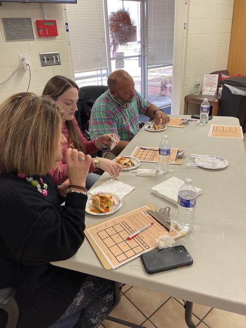 Three people seated at a table eating from paper plates, clipboards and water bottles nearby