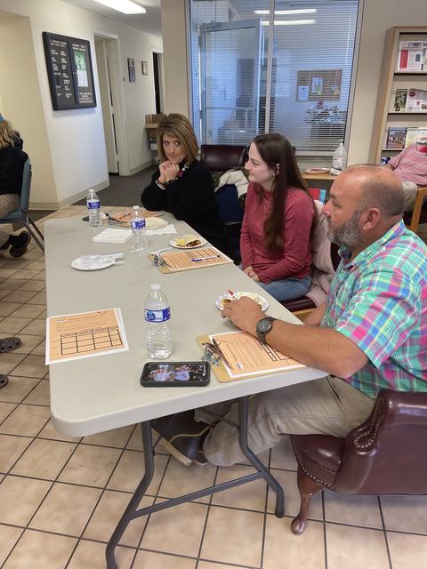 Three adults seated at table with clipboards, water bottles, phone, and snacks