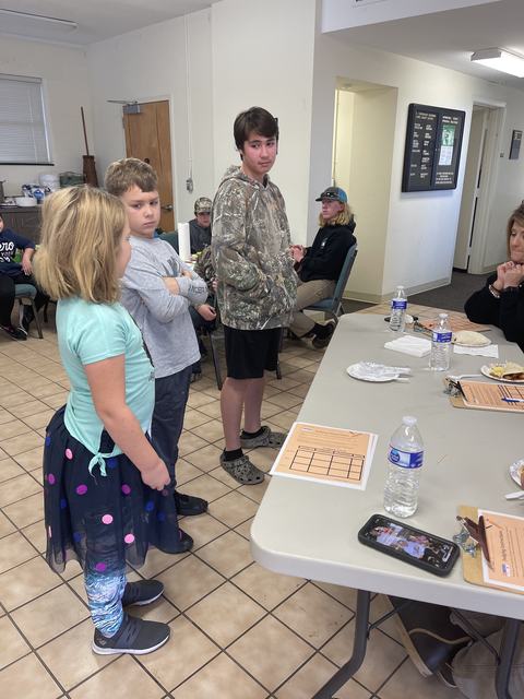 Three children standing by a table with clipboards and water bottles in a meeting room