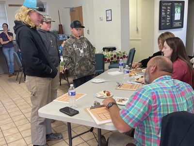 Three boys stand before seated adults at a table with clipboards, water, and snacks