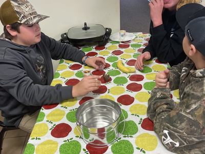 Three boys at a table with apple-patterned cloth, cups, a banana, and a metal bucket.