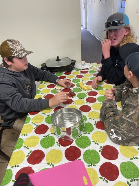 Three boys at a table with apple-patterned cloth, cups, a banana, and a metal bucket.