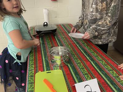Two children at table with green cutting board, orange knife, metal bucket, griddle and card "2"