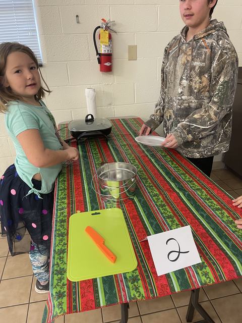 Two children at table with green cutting board, orange knife, metal bucket, griddle and card "2"