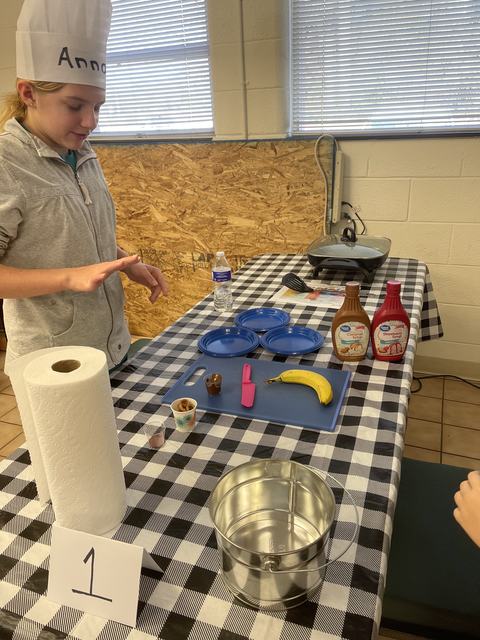 Child in chef hat at table with banana, toppings, syrup bottles, plates, and sign "1".