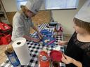 Two children in paper chef hats preparing snacks at a table with pretzels, spreads, and sign "1"