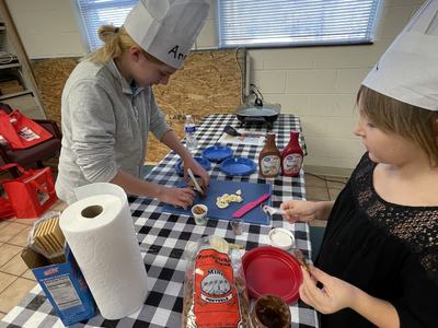 Two children in paper chef hats preparing snacks at a table with pretzels, spreads, and sign "1"