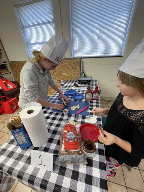 Two children in paper chef hats preparing snacks at a table with pretzels, spreads, and sign "1"