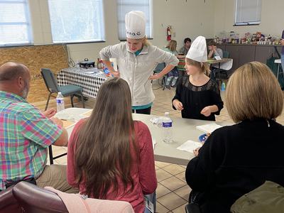 Two girls in chef hats serving food to seated adults at a community room table