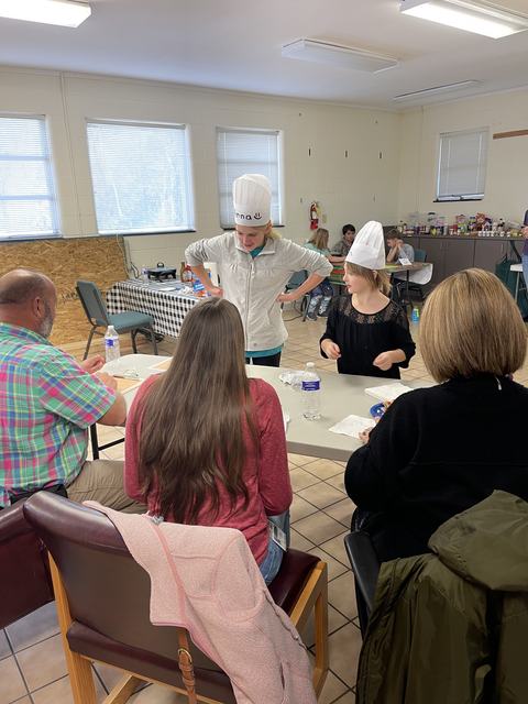Two girls in chef hats serving food to seated adults at a community room table