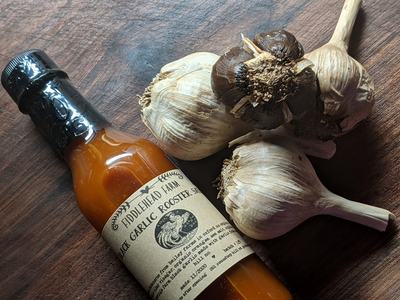 Bottle of Fiddlehead Farm Black Garlic Rooster Sauce beside three garlic bulbs on wooden surface