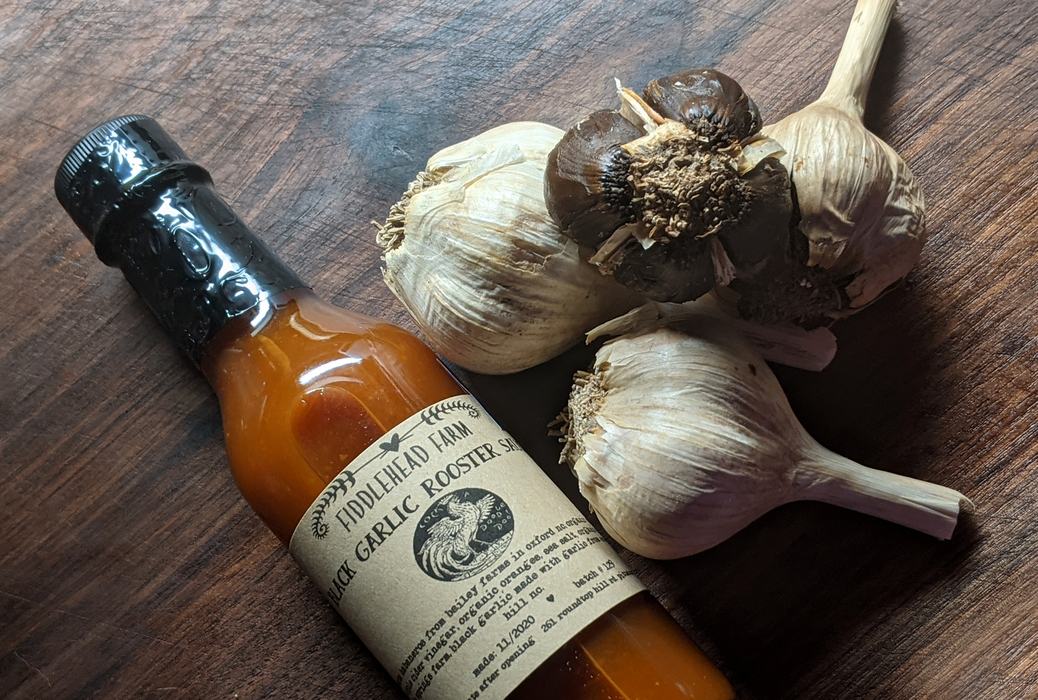 Bottle of Fiddlehead Farm Black Garlic Rooster Sauce beside three garlic bulbs on wooden surface