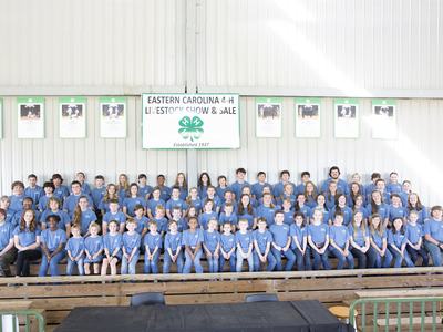 Large group of children and teens in blue shirts on bleachers under sign "Eastern Carolina 4‑H Livestock Show & Sale"
