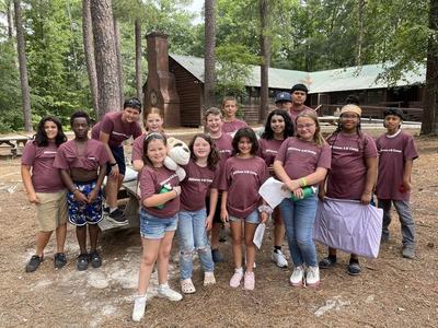 Group of children wearing maroon "Millstone 4-H Camp" shirts in front of wooden cabins in pine woods