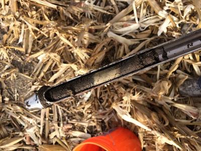 Metal rod split lengthwise showing greasy inner cavity on corn-stalk debris.