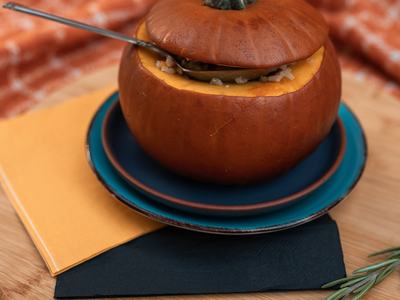Small hollowed pumpkin filled with food and a spoon, on stacked plates and a wooden board.