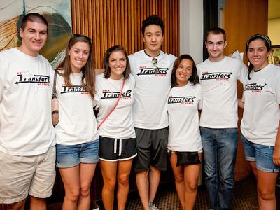 Seven young adults standing indoors wearing white t-shirts reading "Transfers NC State."
