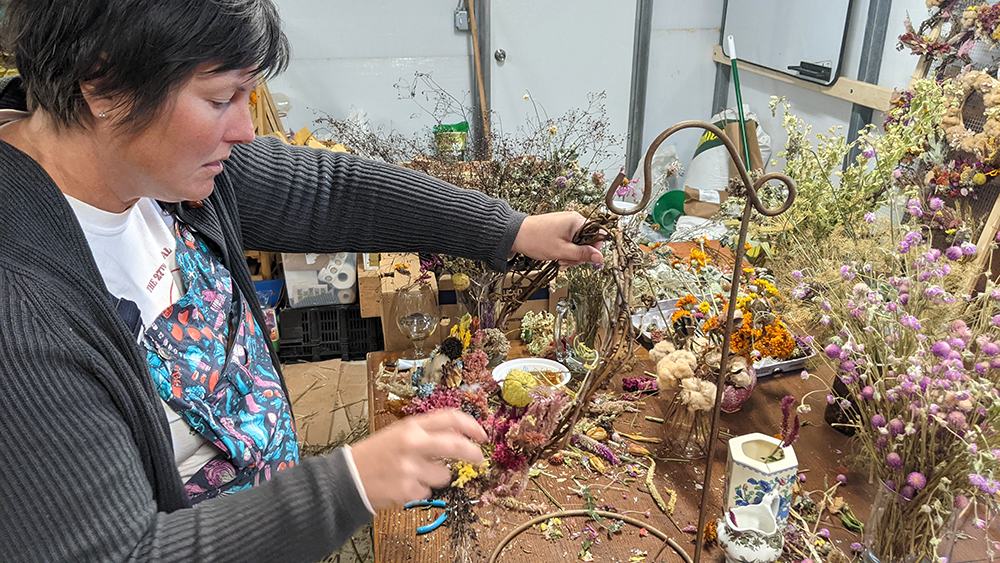 Person assembling dried-flower wreath at a cluttered table with jars and floral tools