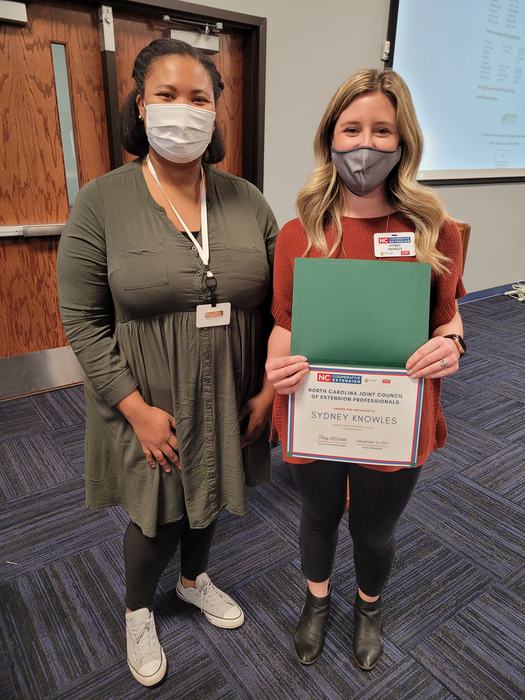 Two masked women standing; right woman holds certificate reading "Sydney Knowles."