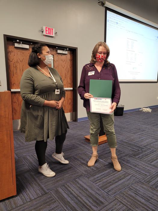 Two masked women standing in a meeting room; right person holds a certificate folder