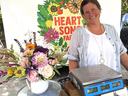 Vendor at market stall with bouquet and scale; sign reads "HEART SONG FARM"