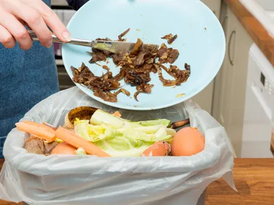 Hand scraping cooked food scraps from blue plate into kitchen trash bin with vegetable peels