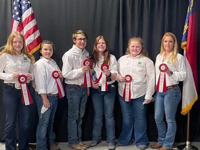 Six 4-H members in white shirts holding red award ribbons with U.S. and state flags behind