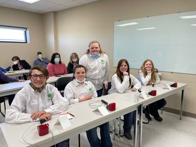 Five youths in white 4-H shirts at classroom tables with red buzzers and seated spectators