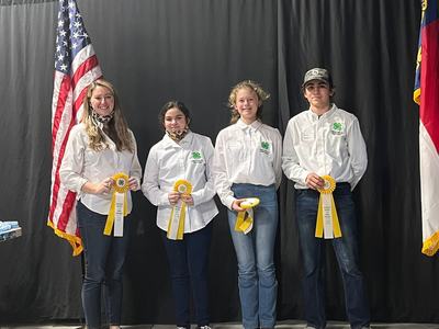 Four youth in white 4-H shirts holding yellow ribbons between U.S. and state flags