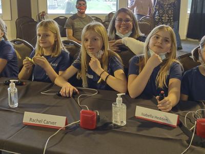 Six girls at a quiz table with buzzers; visible name cards read "Rachel Carver" and "Isabella Hedden."