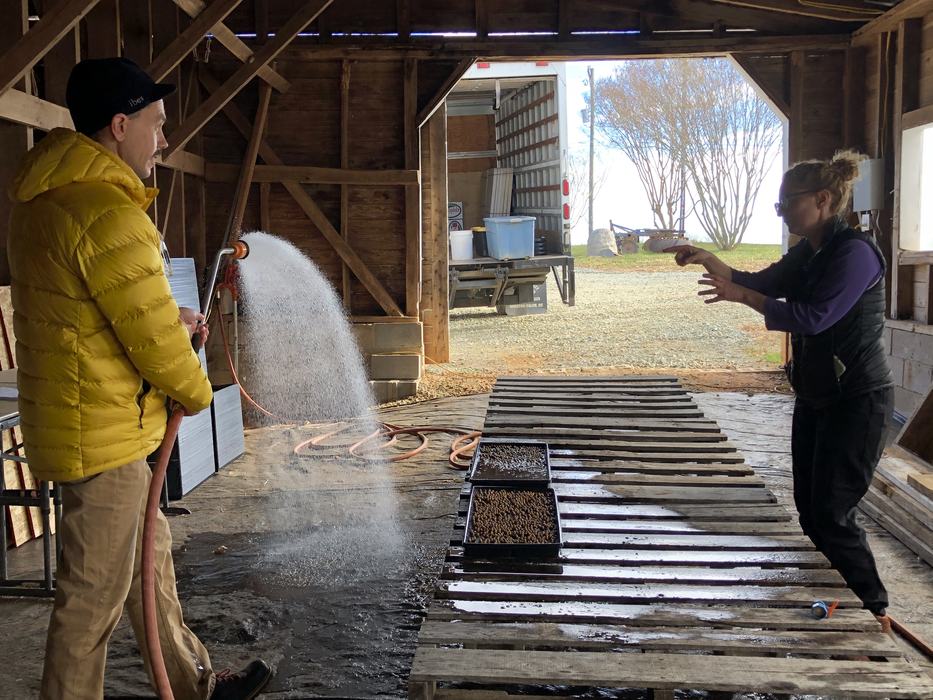 Person in yellow jacket spraying water from a hose toward another person across wooden pallets
