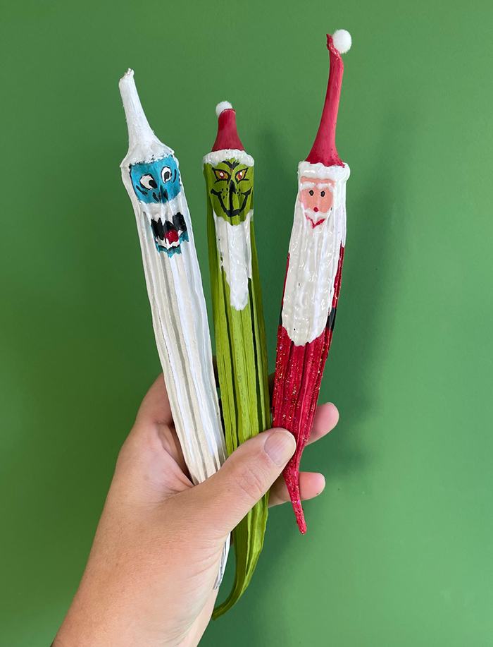 Hand holding three painted dried pods with faces: red Santa, green grinning figure, blue-faced figure